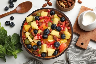 Delicious fruit salad in bowl, berries, fresh mint and nuts on white marble table, flat lay