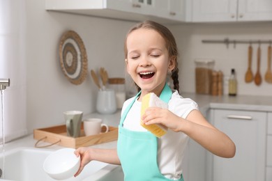 Photo of Cute little girl with sponge singing while washing dishes in kitchen