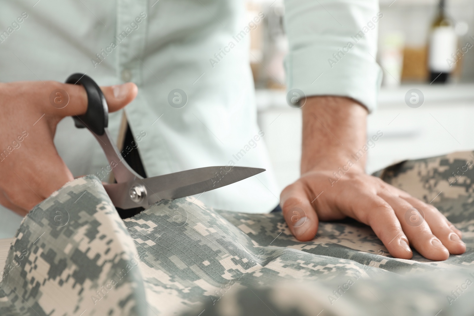 Photo of Professional tailor cutting camouflage fabric with scissors in workshop, closeup