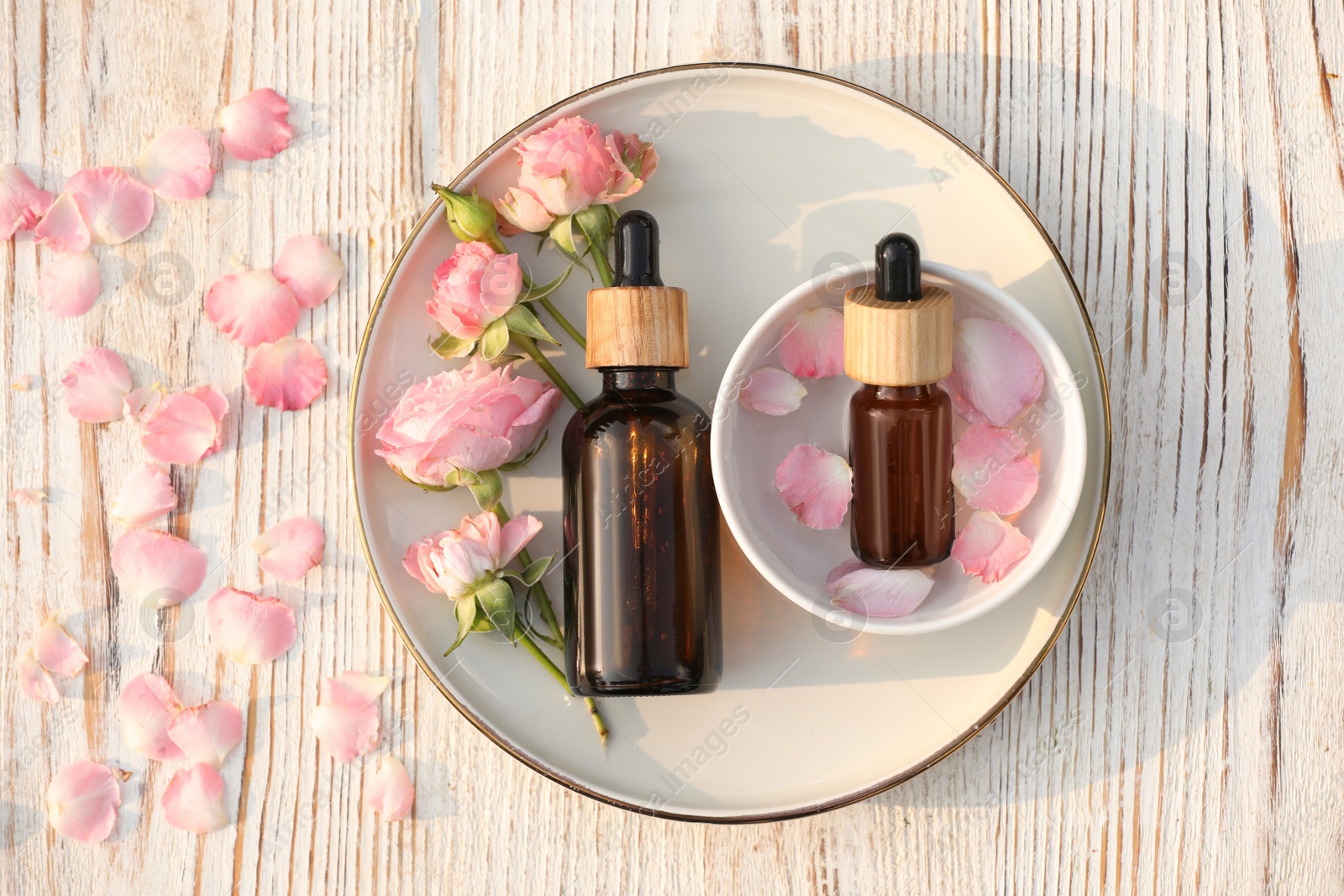 Photo of Bottles of rose essential oil and flowers on white wooden table, flat lay