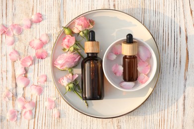 Bottles of rose essential oil and flowers on white wooden table, flat lay