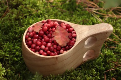 Photo of Many tasty ripe lingonberries and autumn leaves in wooden cup outdoors, closeup