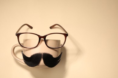 Artificial moustache, cup and glasses on beige background, top view. Space for text