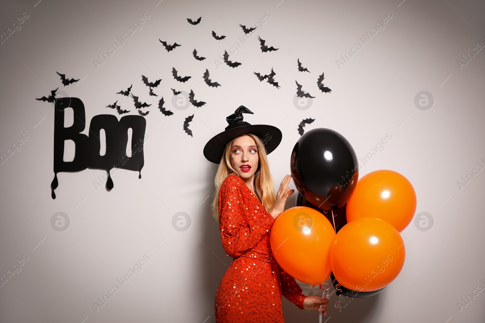 Photo of Woman in witch hat with balloons posing near white wall decorated for Halloween