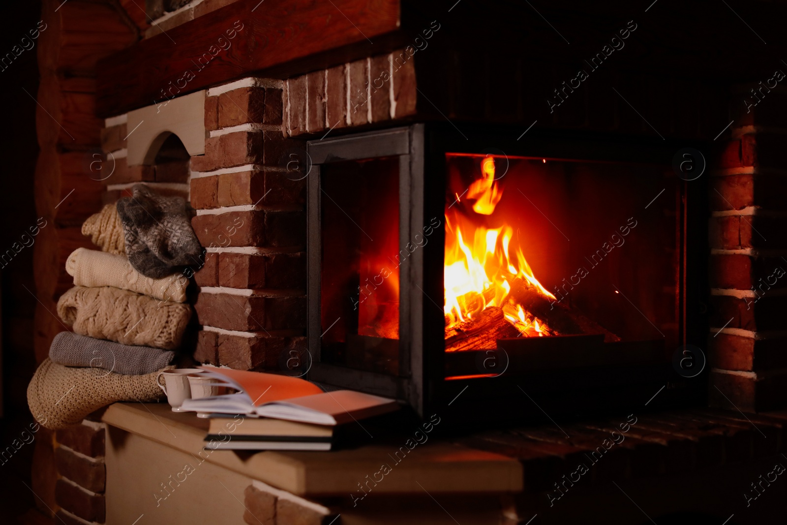 Photo of Stack of knitwear near fireplace with burning wood indoors. Winter vacation