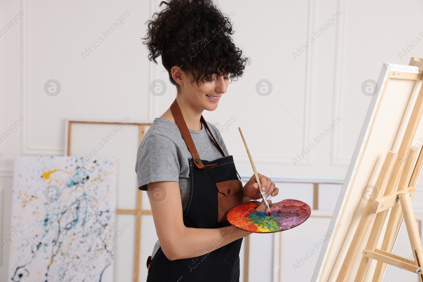 Photo of Young woman mixing paints on palette with brush near easel in studio