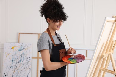 Young woman mixing paints on palette with brush near easel in studio