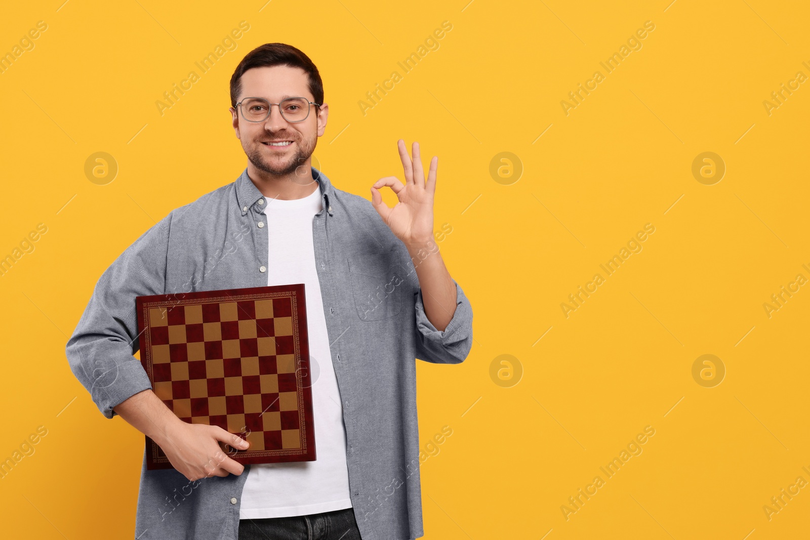 Photo of Smiling man holding chessboard and showing OK gesture on orange background. Space for text