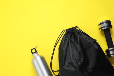 Black drawstring bag, thermo bottle and dumbbell on yellow background, flat lay. Space for text