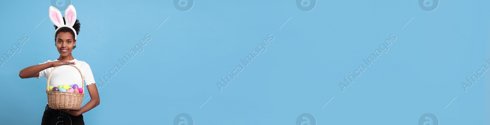 Image of African American woman with bunny ears holding basket full of Easter eggs on light blue background, space for text. Banner design