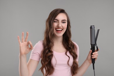 Beautiful young woman with curling hair iron showing OK gesture on grey background