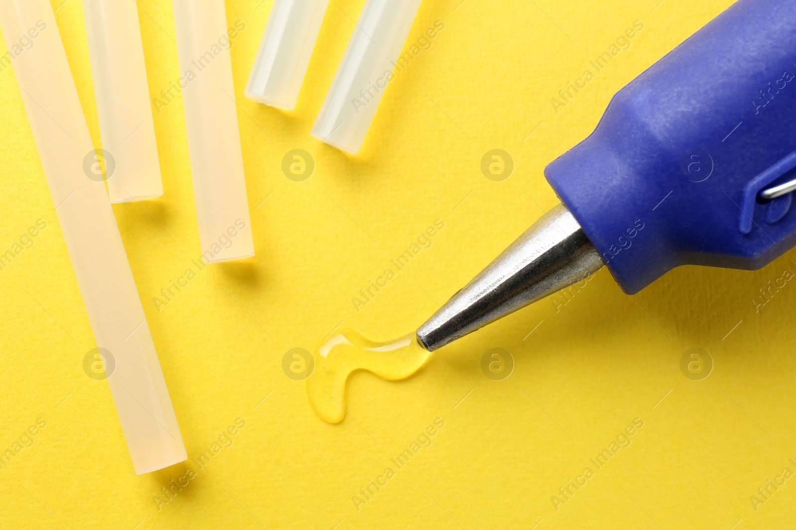 Photo of Melted glue dripping out of hot gun nozzle near sticks on yellow background, closeup