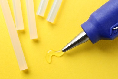 Photo of Melted glue dripping out of hot gun nozzle near sticks on yellow background, closeup