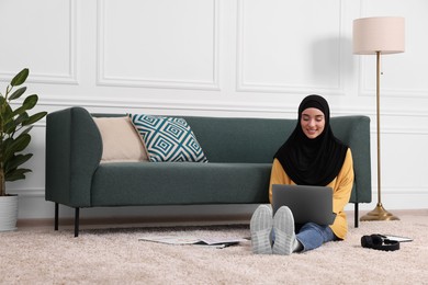 Photo of Muslim woman in hijab using laptop on floor near sofa in room. Space for text