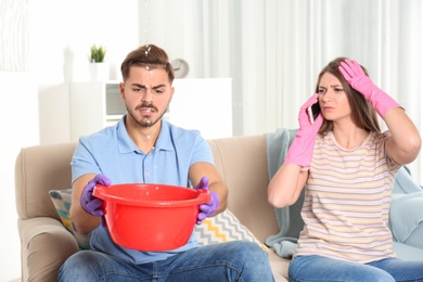 Worried young man collecting water leakage from ceiling while his wife calling plumber at home