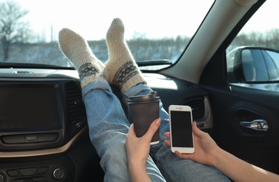 Photo of Young woman with phone in warm socks holding her legs on car dashboard and drinking coffee. Cozy atmosphere