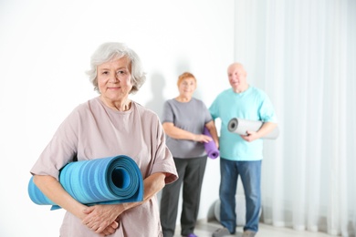 Elderly woman with yoga mat indoors. Space for text