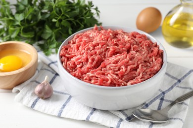 Raw ground meat in bowl, garlic, egg, parsley and spoon on white table, closeup