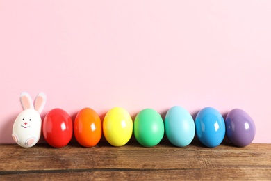 Photo of Bright Easter eggs and white one as bunny on wooden table against pink background, space for text