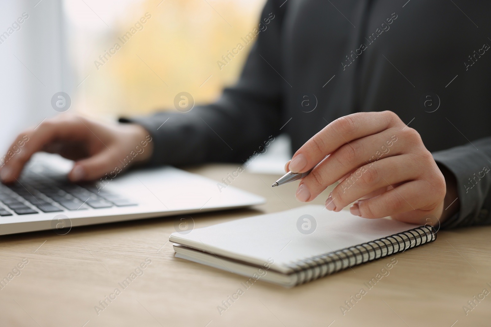 Photo of Woman with notebook and pen working on laptop at wooden table, closeup. Electronic document management