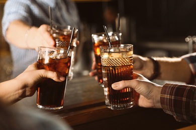 Photo of Group of friends clinking glasses with cola at table indoors, closeup