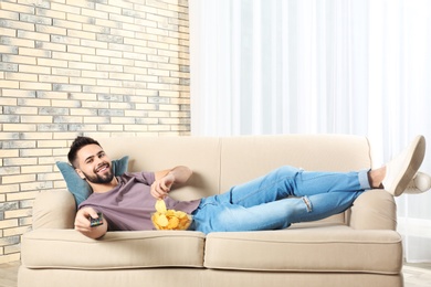 Young man with remote control and bowl of chips watching TV on sofa at home