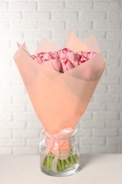 Bouquet of beautiful pink tulips in vase on white table near brick wall