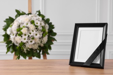 Photo frame with black ribbon on table and wreath of flowers near white wall indoors, space for text. Funeral attributes