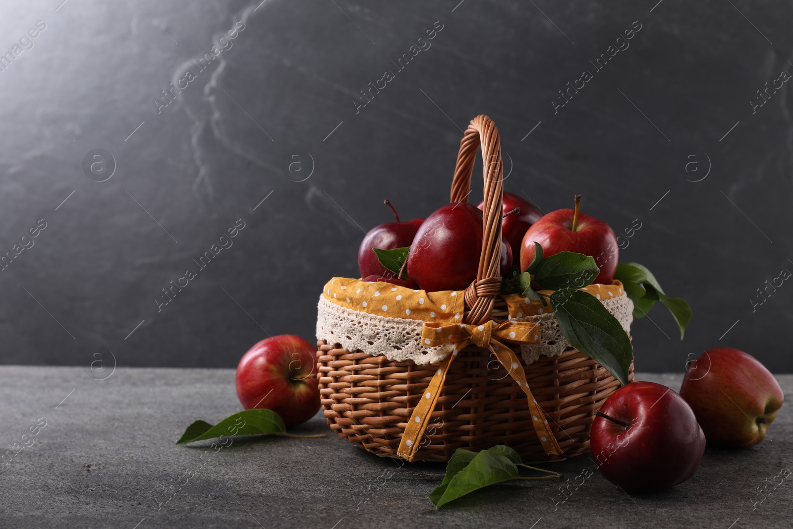 Photo of Ripe red apples and leaves in wicker basket on dark grey table. Space for text