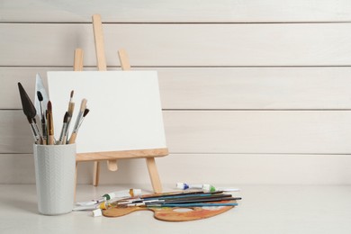 Photo of Easel with blank canvas, brushes, paints and palette on white wooden table. Space for text