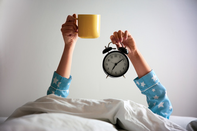 Woman with cup and alarm clock lying in bed, closeup. Morning time