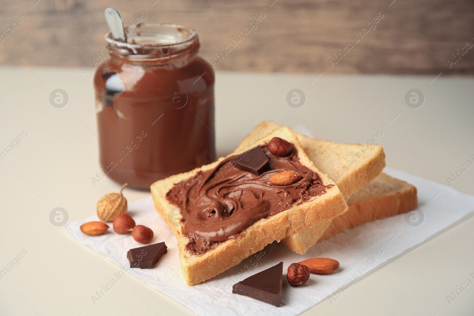 Photo of Tasty toast with chocolate paste and nuts near jar of cream on light table