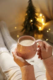 Photo of Woman holding cup with hot drink near festive lights at home, closeup