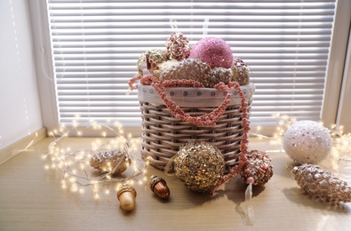 Basket with beautiful Christmas tree baubles and fairy lights on window sill indoors