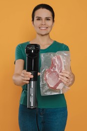 Photo of Beautiful young woman holding sous vide cooker and meat in vacuum pack on orange background