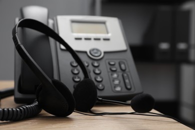 Photo of Modern headset near stationary phone on wooden desk in office, closeup. Hotline service