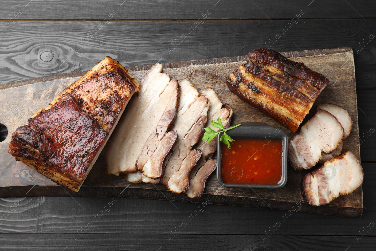 Photo of Pieces of baked pork belly served with sauce and parsley on black wooden table, top view