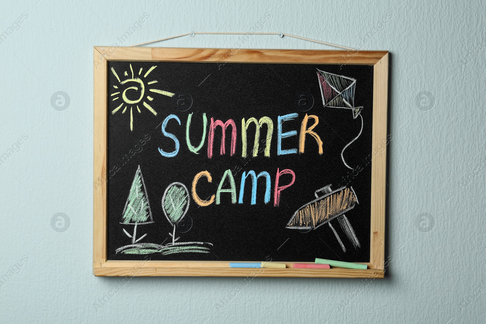 Photo of Small blackboard with text SUMMER CAMP, drawings and chalk sticks hanging on color wall