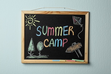 Photo of Small blackboard with text SUMMER CAMP, drawings and chalk sticks hanging on color wall