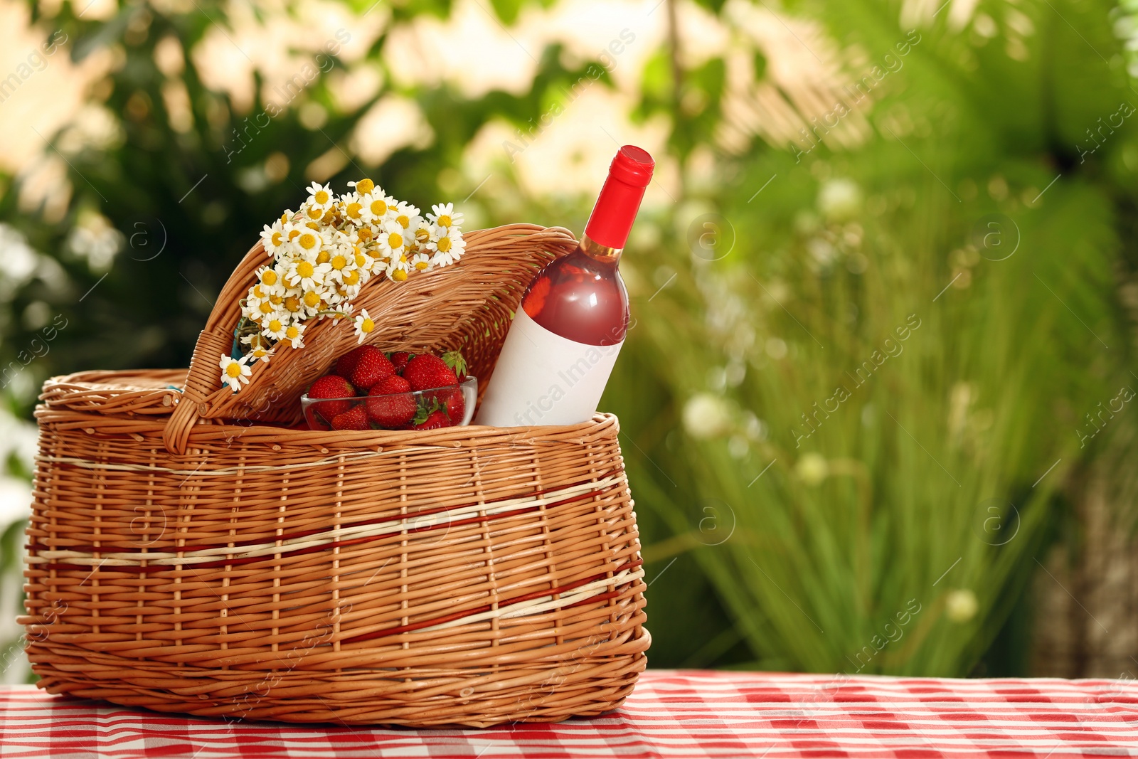 Photo of Picnic basket with wine, strawberries and flowers on checkered tablecloth against blurred background, space for text
