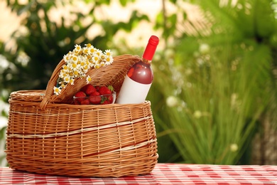 Picnic basket with wine, strawberries and flowers on checkered tablecloth against blurred background, space for text