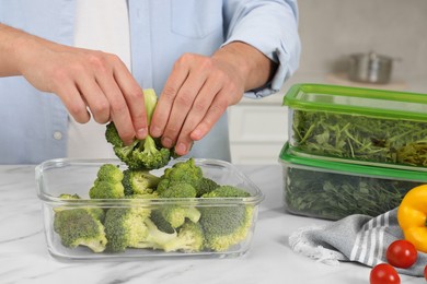 Man putting broccoli into glass container at white marble table in kitchen, closeup. Food storage