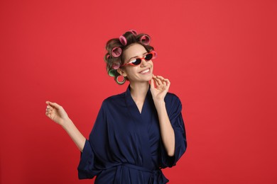 Photo of Beautiful young woman in silk bathrobe with hair curlers and sunglasses on red background