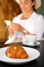Woman sitting near tasty pastry and cup of aromatic coffee at black table in cafeteria, focus on croissant