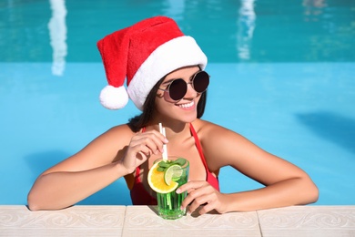 Photo of Young woman wearing Santa Claus hat with refreshing drink in swimming pool. Christmas vacation