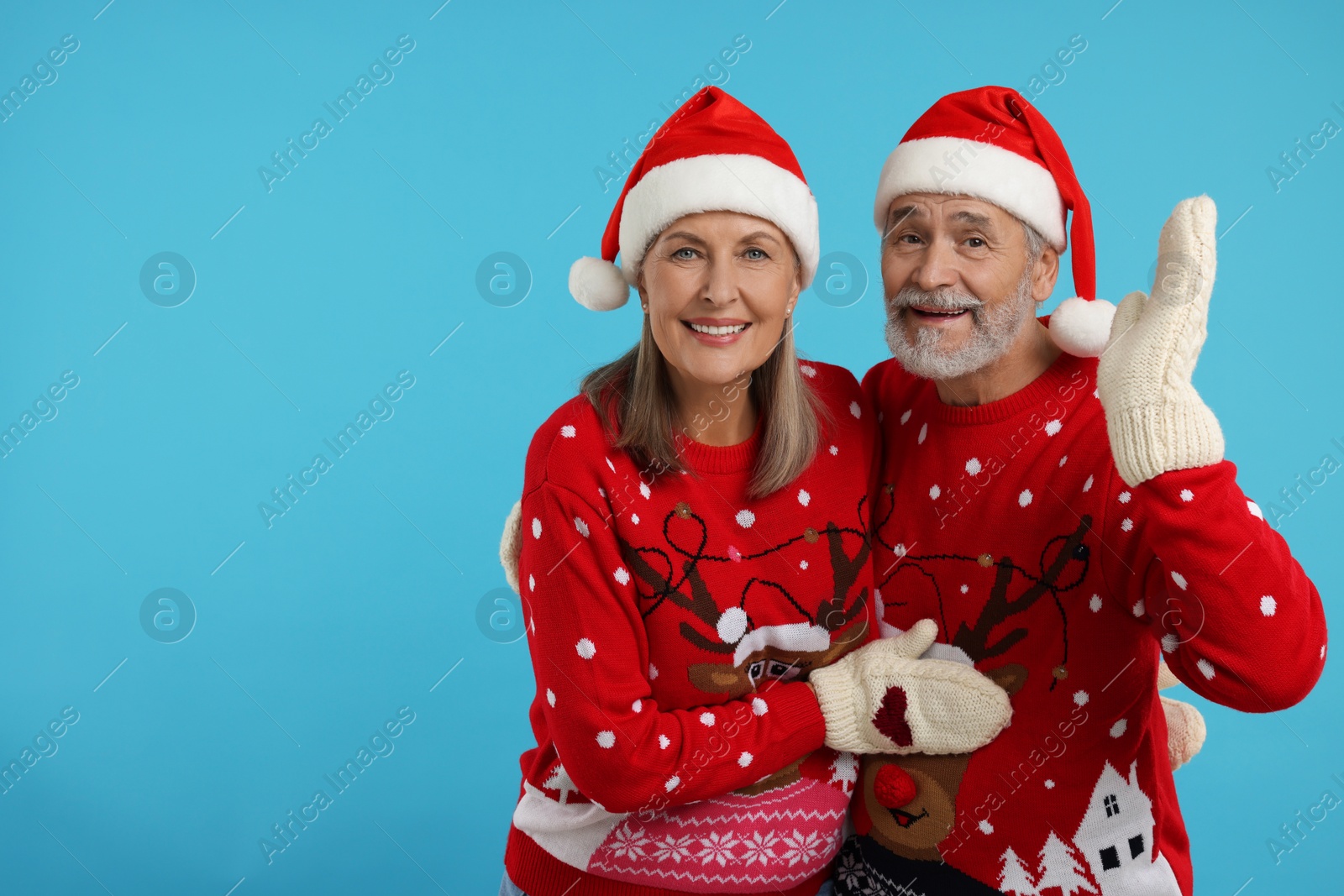 Photo of Happy senior couple in Christmas sweaters, Santa hats and knitted mittens on light blue background. Space for text