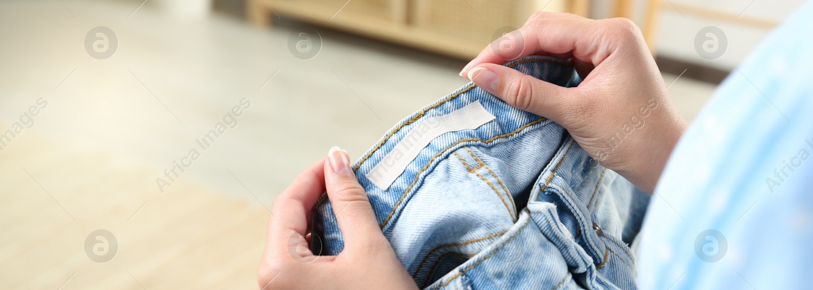 Image of Woman holding jeans with blank clothing label at home, closeup. Banner design