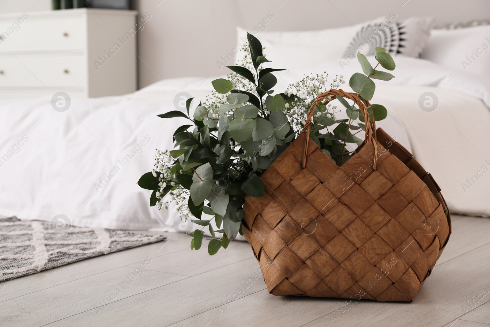 Photo of Stylish wicker basket with fresh eucalyptus branches and flowers on floor in bedroom