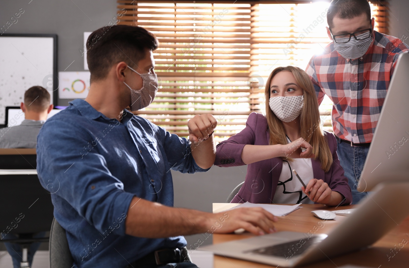 Photo of Coworkers with protective masks making elbow bump in office. Informal greeting during COVID-19 pandemic