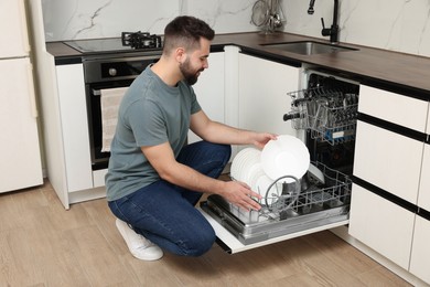 Smiling man loading dishwasher with plates in kitchen